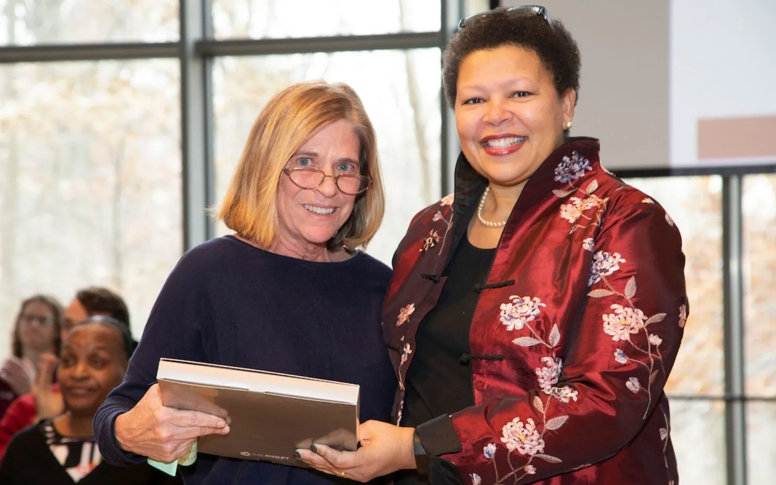 Two women stand side by side and smile at the camera. The one on the right wears a black top and has shoulder length blonde hair and glasses. The one on the left has short dark hair and wears a deep red patterned silk jacket.
