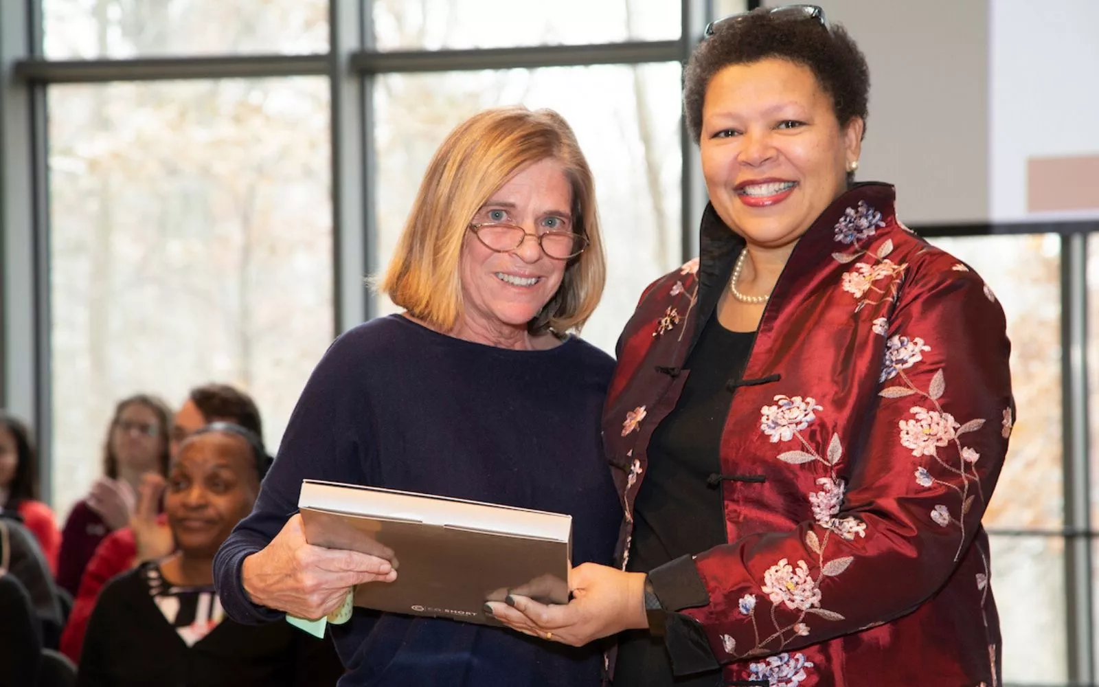 A smiling woman with blonde hair and glasses stands next to a smiling woman with short hair wearing a patterned red silk jacket, handing her an award.