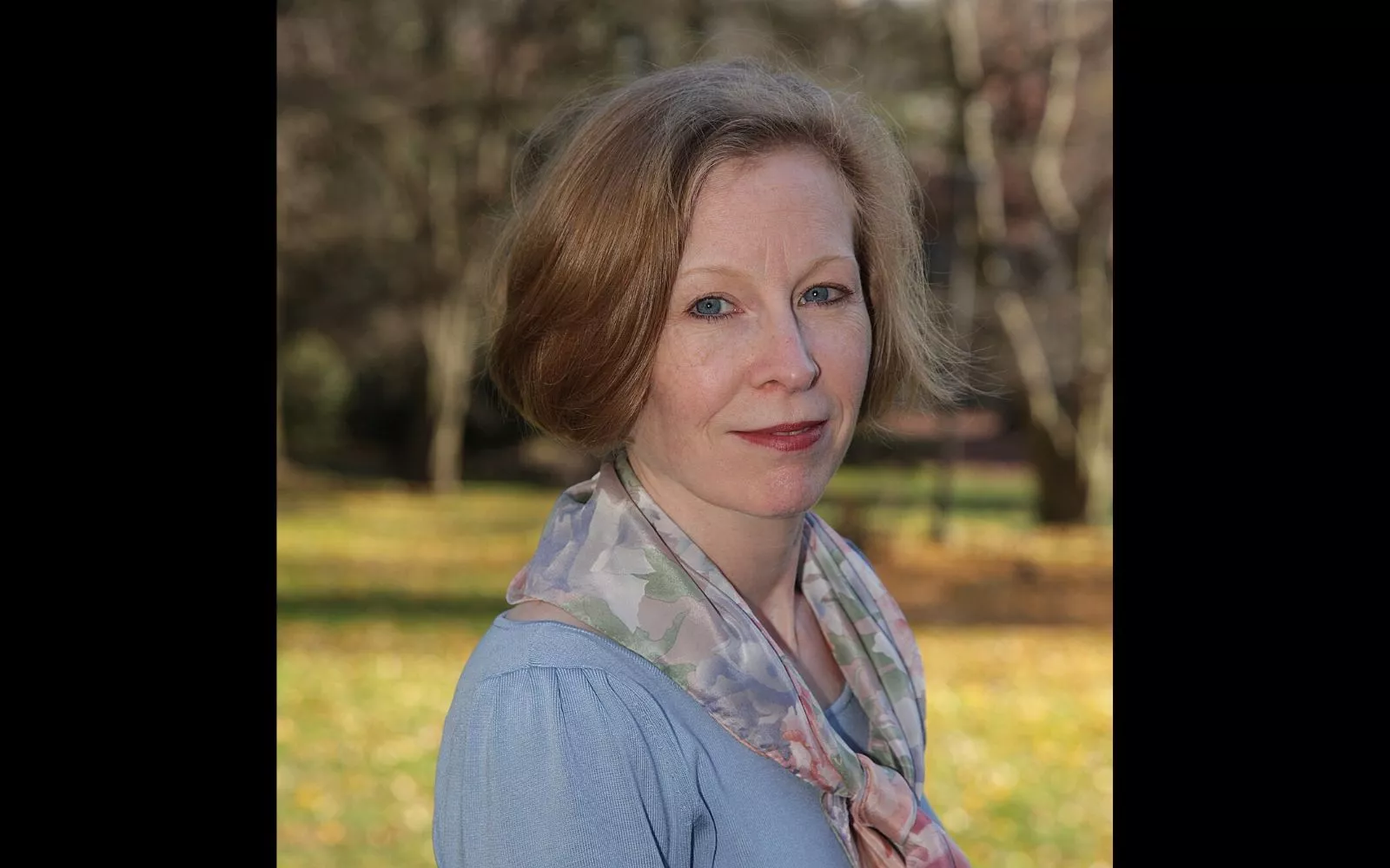 Headshot of woman with short brown hair wearing a light blue top and silk scarf.