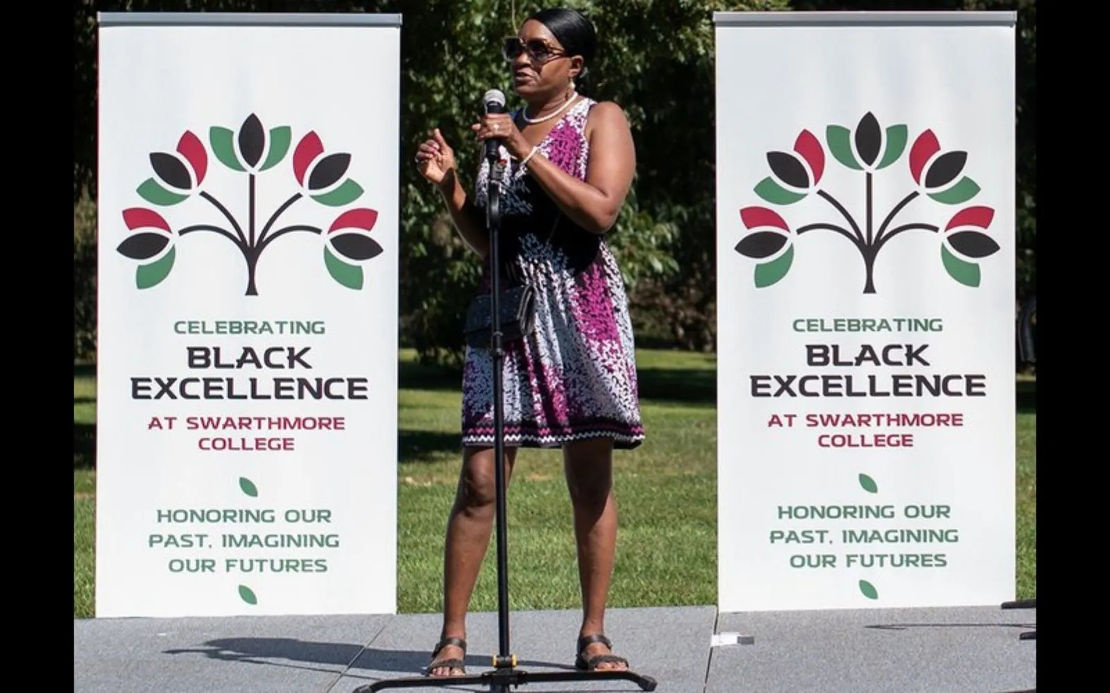 A woman with short hair, wearing a patterned dress and surrounded by two banners with the words "Black Excellence at Swarthmore" speaks into a microphone.