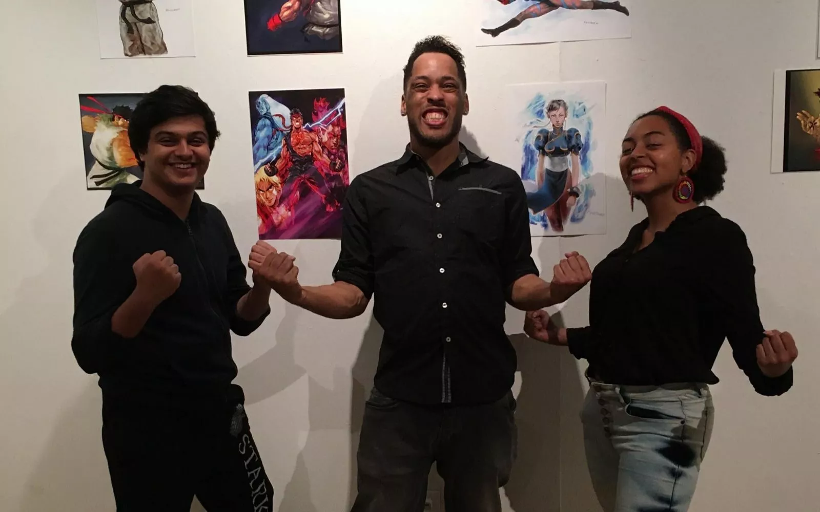Three people pose, grinning, in front of a wall of art. The man in the center is Steve Mack, who wears a dark button down shirt and pants and has a short beard and short hair 