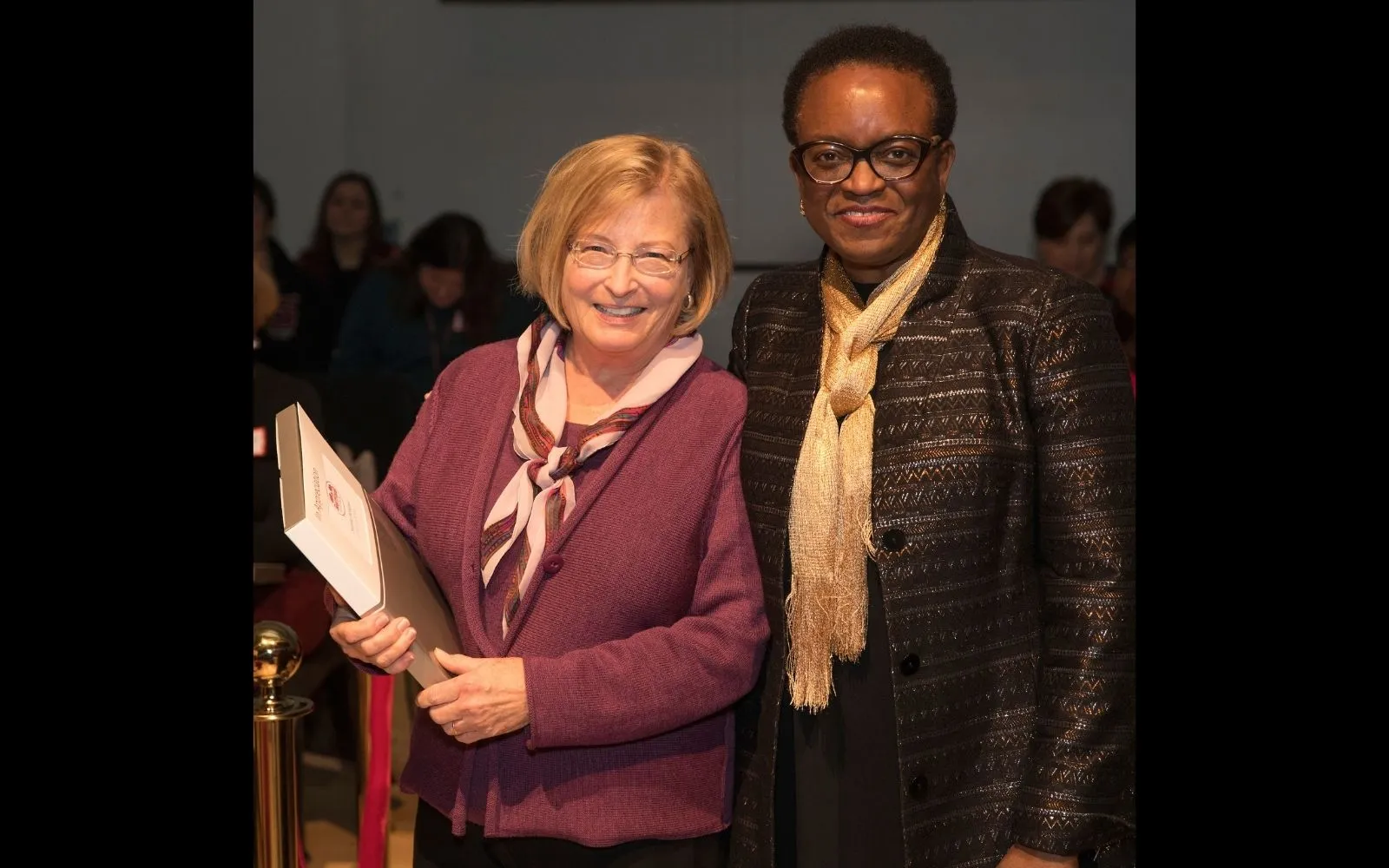 A woman with short blonde hair wearing a purple top and pink scarf stands next to a woman with close cropped hair, glasses, and a brown jacket. Both are smiling.