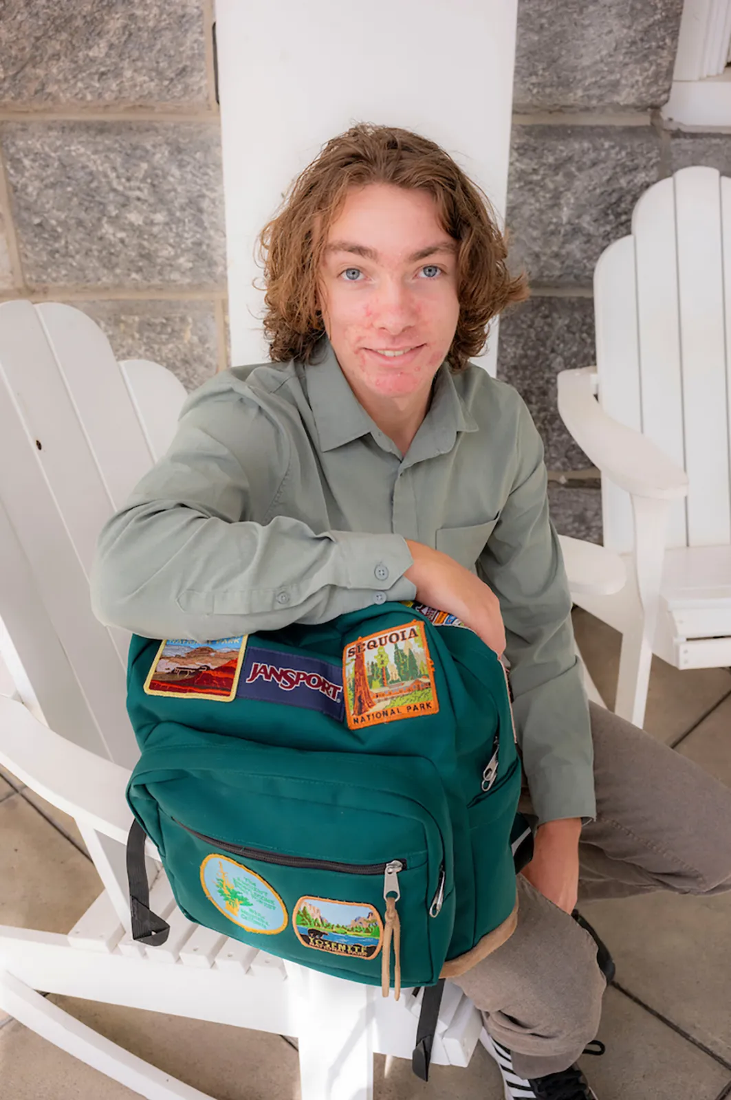 Student sitting in white Adirondack chair holds green hiking backpack