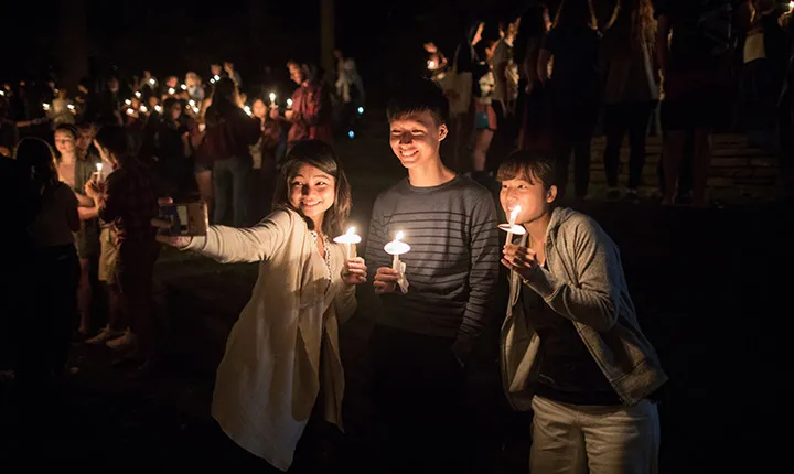 Three students take a selfie together while holding candles outside at night