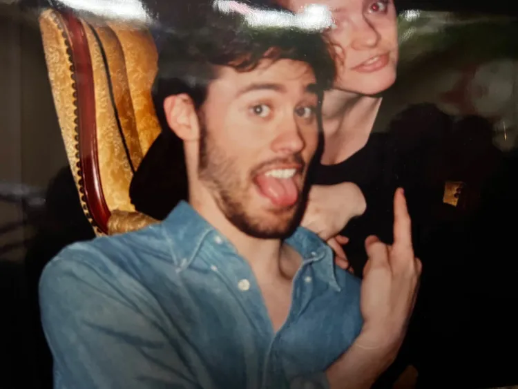A photo of Dito Van Reigersberg wearing a blue chambray shirt and pointing up. He's sticking his tounge out
