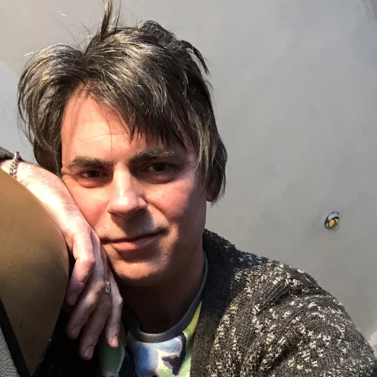 Adriano Shaplin leans against the arm of a chair and smiles at the camera.