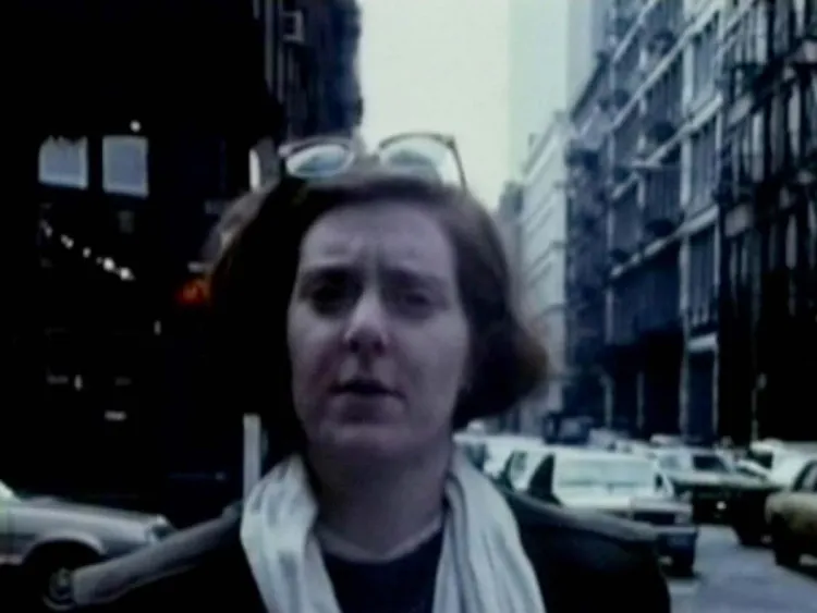 Still from Superstar A Karen Carpenter Story featuring a woman with a dark brown bob haircut and a light scarf standing on a street in a city with cards and tall building behind her. She is wearing sunglasses on the top of her head
