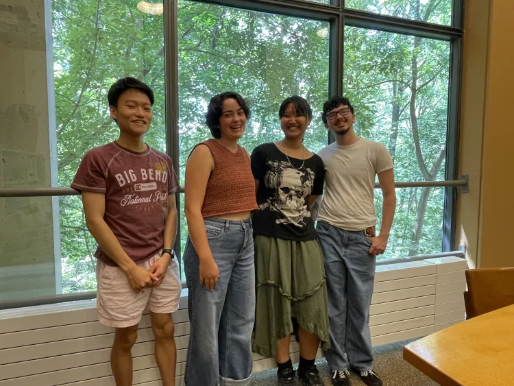 Four students smiling while standing in front of the Underhill Seminar Room window overlooking Crum Woods