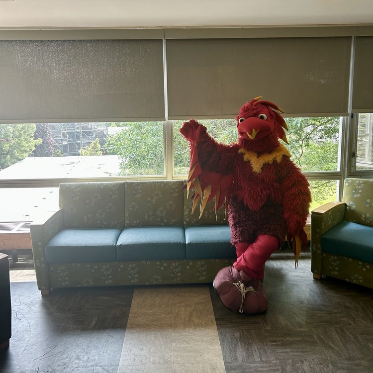 A photo of Swarthmore mascot Phineas the Phoenix in a lounge in Willets Hall sitting on the arm of a sofa waving at the camera.