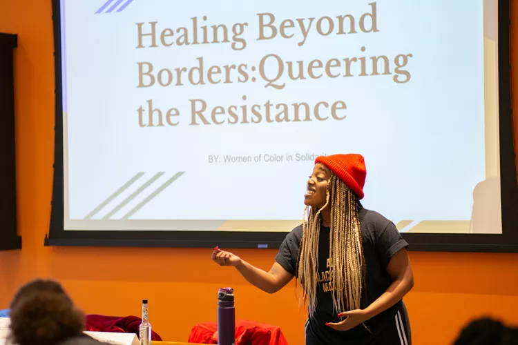 Student in front of screen that says "Healing Beyond Borders: Queering the Resistance"