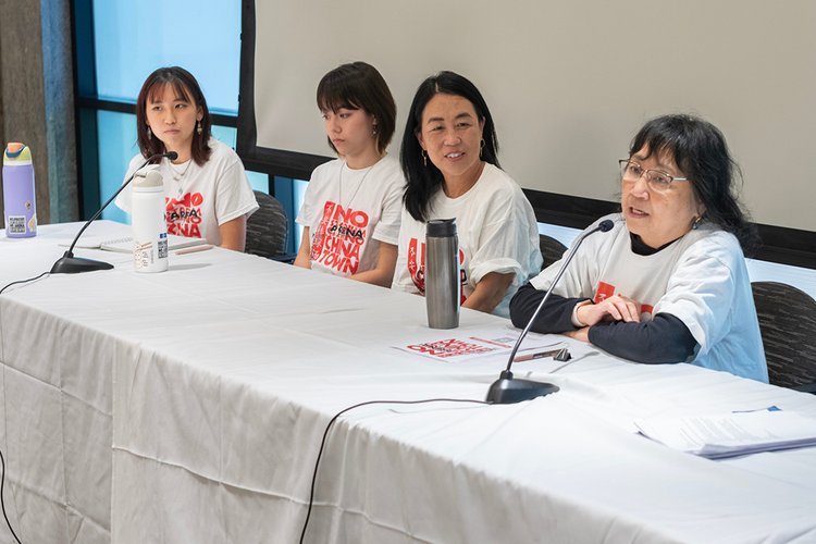 Four women seated at table wearing white shirts that read "No Arena in Chinatown" in red lettering.