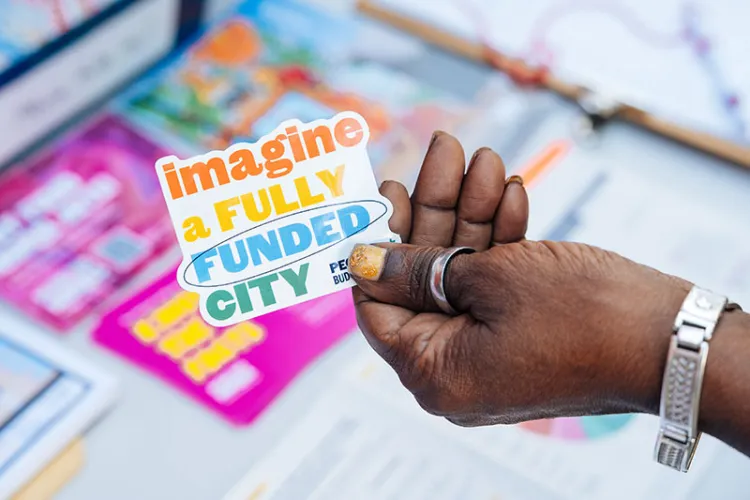 Person holds sticker that says "imagine a fully funded city"