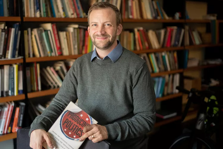 Daniel Laurison sits in office while holding book entitled "Producing Politics"