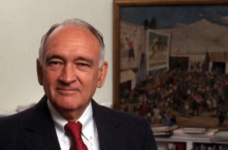 William Spock seated wearing red tie in front of painting in background