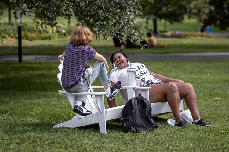 Two students sit outside in white adirondack chairs
