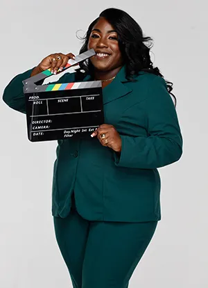 Maziha Cooper wearing green suit in front of white background holds blank film slate.