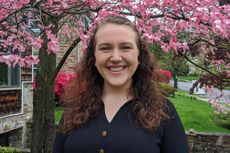 Shay Downey '22 smiling in front of tree with pink flowers and green grass.