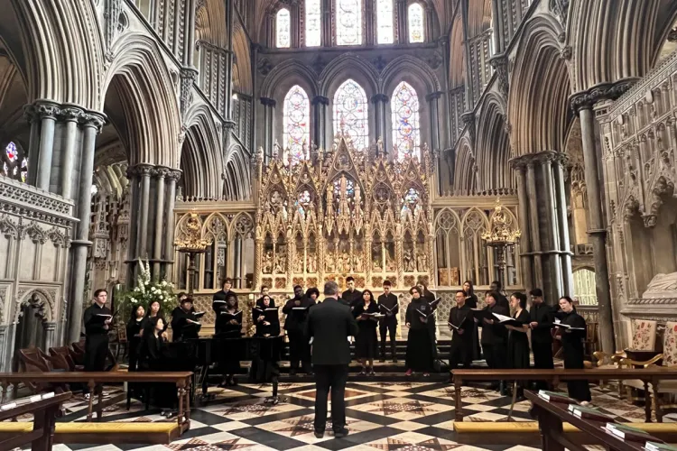 Garnet Singers in two rows, dressed in concert black attire and holding folders, singing in front of a large, intricate backdrop and stained glass windows]