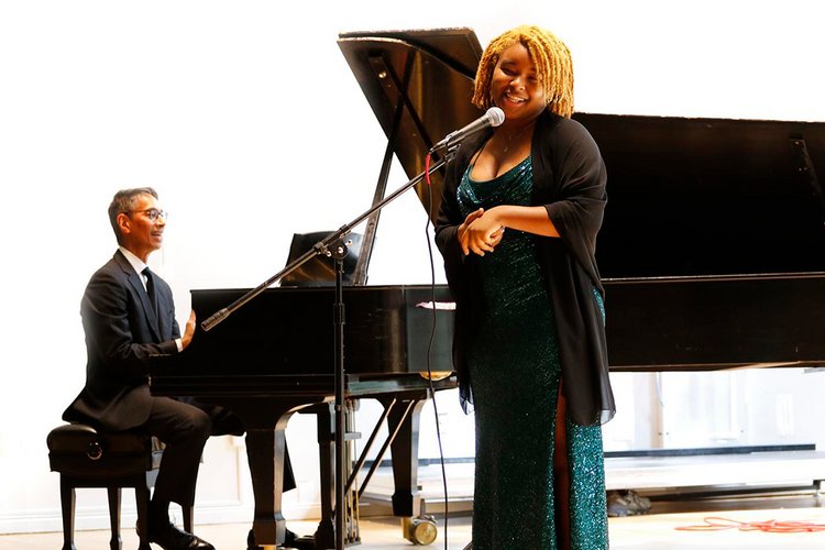 Woman in dress sings at microphone while man accompanies her on piano