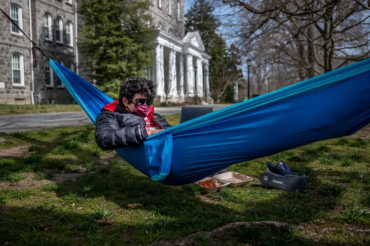 Student wearing mask and sunglasses uses laptop in blue hammock.