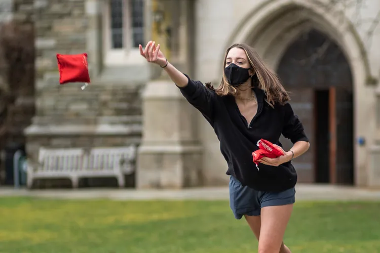 Student wearing mask tosses red bean bag outdoors.