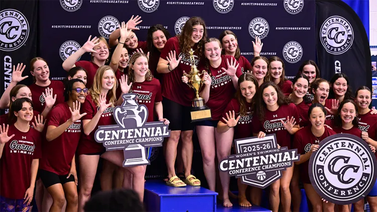 Women's swimming team celebrates conference title with group photo on podium and holding trophy while holding up five fingers.
