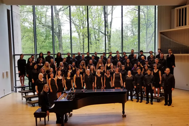 Members of chorus chorus stand on risers in front of large glass window