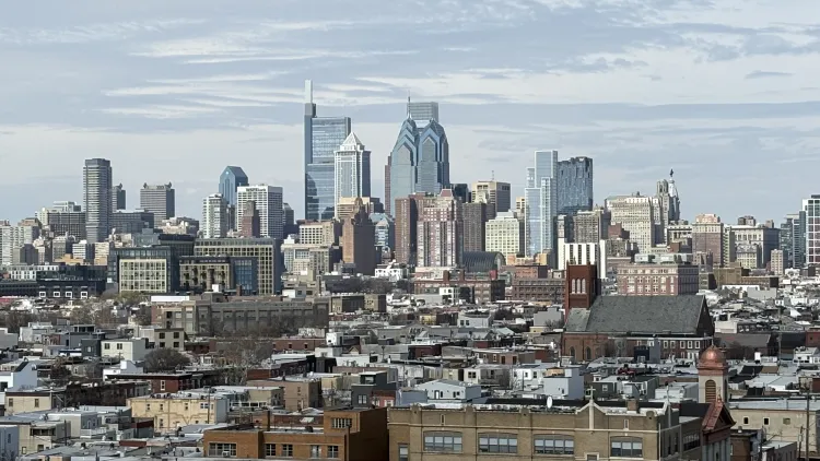 Image of view of Philly skyline from atop the Bok Building in South Philly, showing skyscrapers in the background, with low rise buildings and houses in the foreground, under a blue sky. Photo taken by Tanya Hoard.