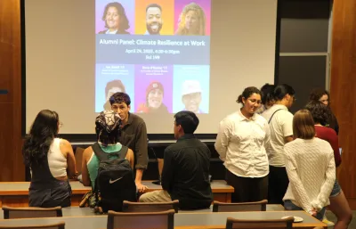 Image of students and alumni chatting, with poster in background showing six alumni who were on the panel