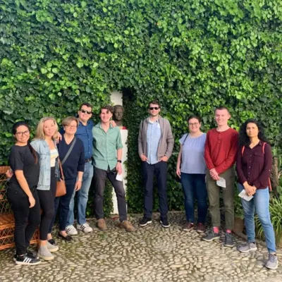 Group of students and faculty stand in semicircle in front of a tall garden hedge