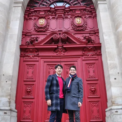 Two male students stand in front of a decorate red exterior door wearing overcoats