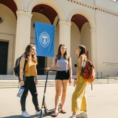 Group of 3 females students stand in sunny quad with a scooter