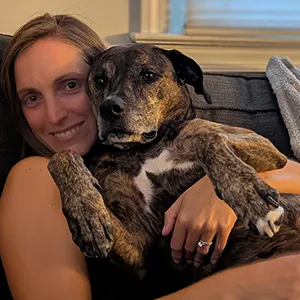 Kathryn Melvin seated comfortably on a sofa and cuddling a very cute dark short haired dog