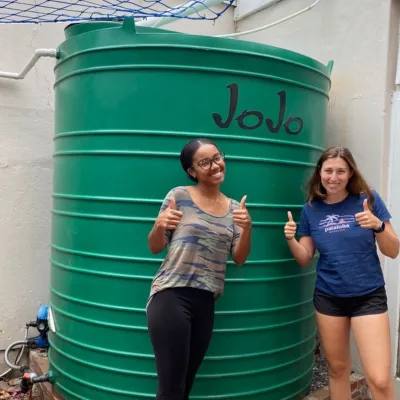 Two students stand with thumbs up in front of a water storage tank
