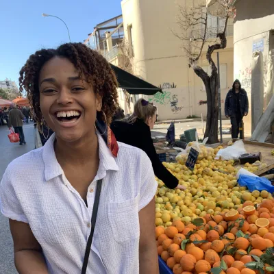 Student smiles widely in front of a produce stall in Greece.