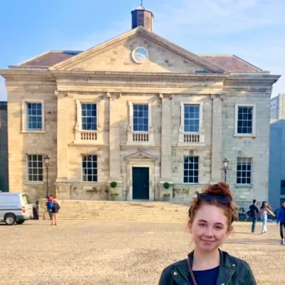 Female student with red hair stands on cobblestone walkway in front of large stone academic building