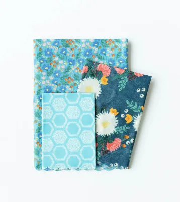 This image depicts a stack of three folded beeswax wraps, which are cotton rectangles coated in beeswax, in three different sizes and designs. All are bright blue floral themes. 