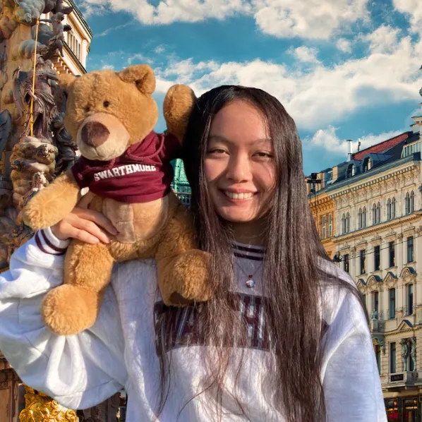 Student with teddy bear smiles in sunny plaza.