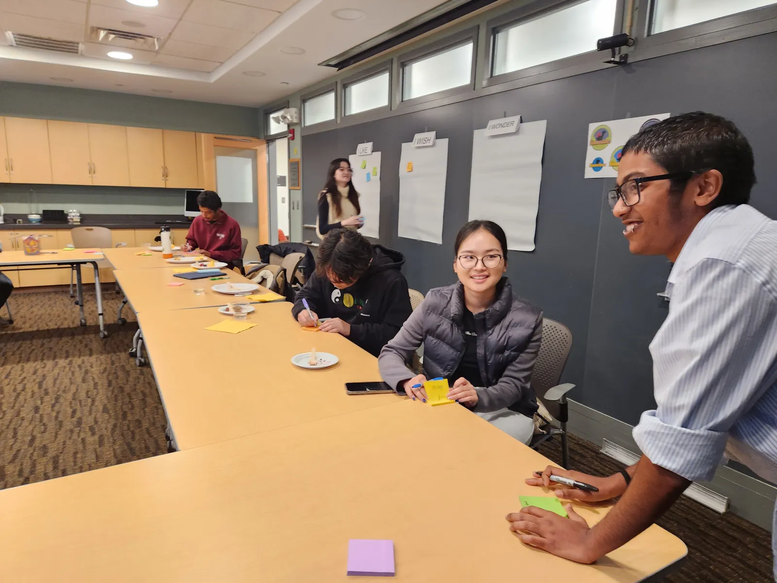 Students smiles, eat, and work at a long table as blackboard behind them is being filled with sticky notes from other students