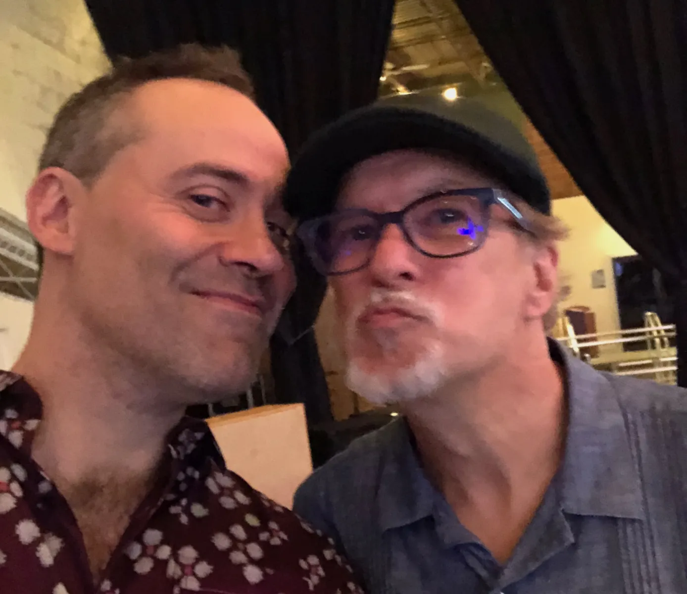 Two white male identified individuals with their faces close to each other. Dito, on the left, wears a patterned shirt, only half his face is visible with an inscrutable smile. Allen, on the right, wears dark rimmed glasses and a pageboy hat. He is making a goofy face next to Dito. 