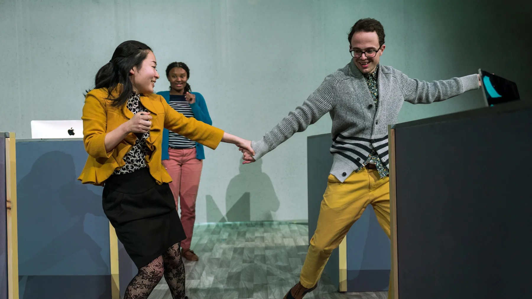 Two people in business casual attire dance in an office while a third watches on from the background in joy