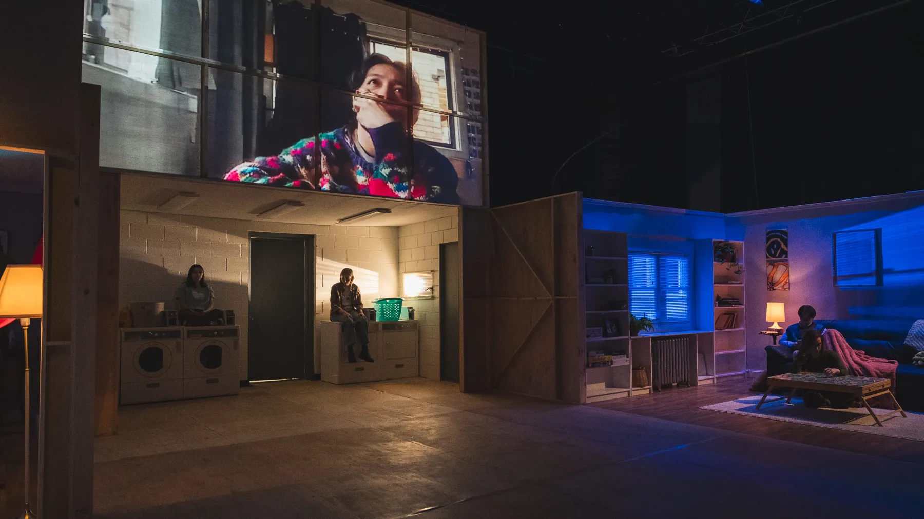 An image from this light says i love you, an Honors Thesis in Directing. A laundry room is wash in warm light, while two individuals sit atop the machines. Above the laundry room is a still from a web-conferencing call featuring an individual in a green and red sweater. They have their hand over their mouth. Across from the laundry room is a living room illuminated in cool blue light. A solitary figure sits on the couch under a blanket.