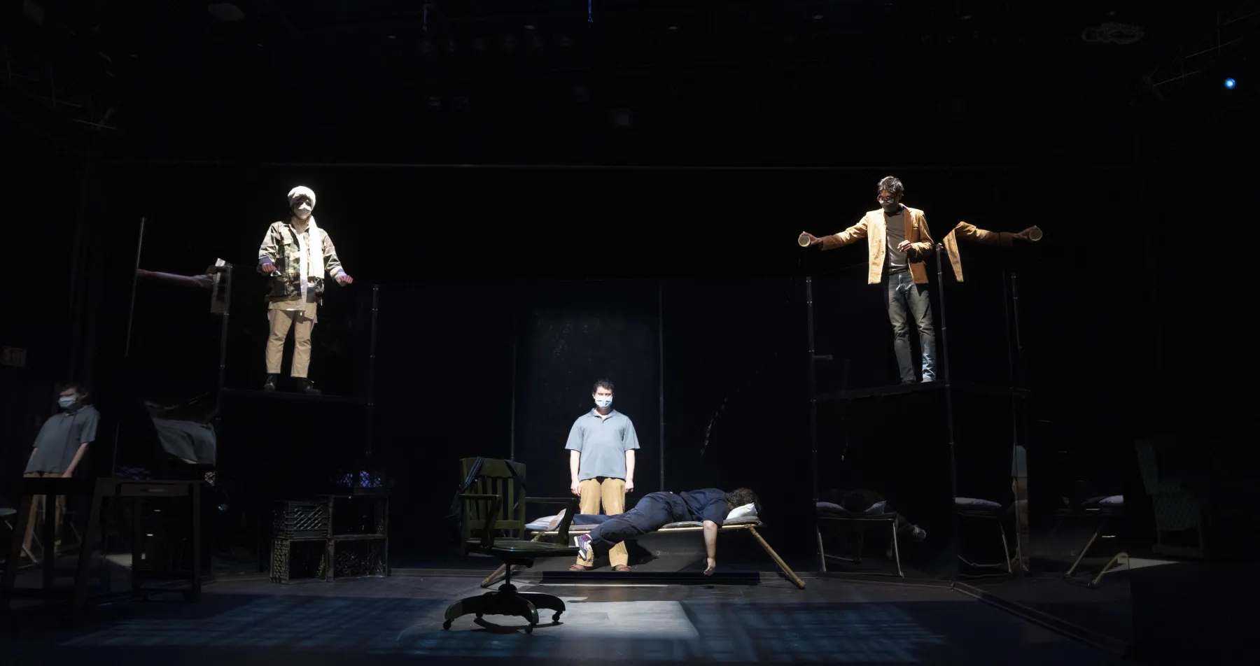 A dark stage with spotlights on four actors - one lying face down and three standing above. 
