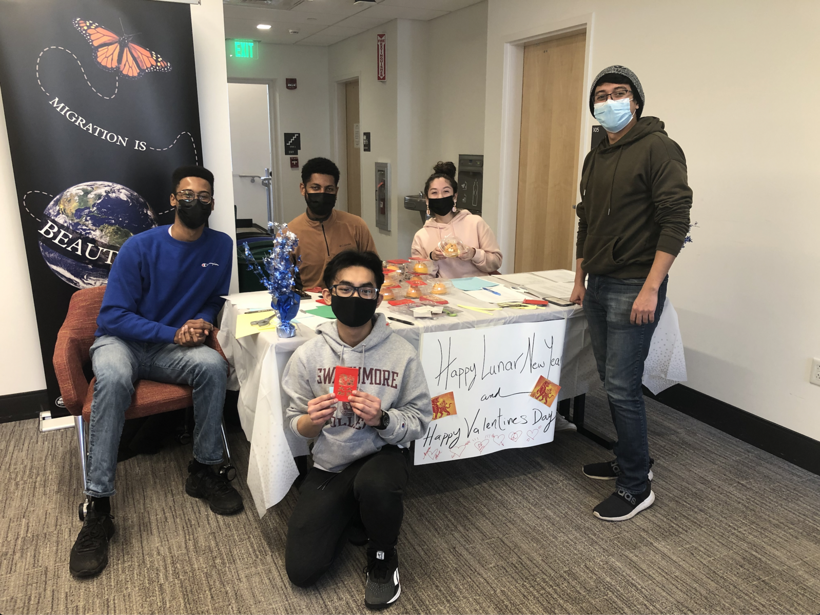 Masked IC Interns stand around a table, indoors, with a sign that says "Lunar New Year." They are crafting good luck messages to hand out, along with pastries, for the new year.