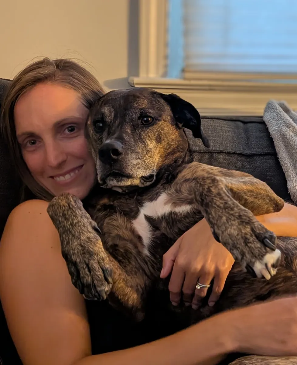 Kathryn Melvin seated comfortably on a sofa and cuddling a very cute dark haired dog
