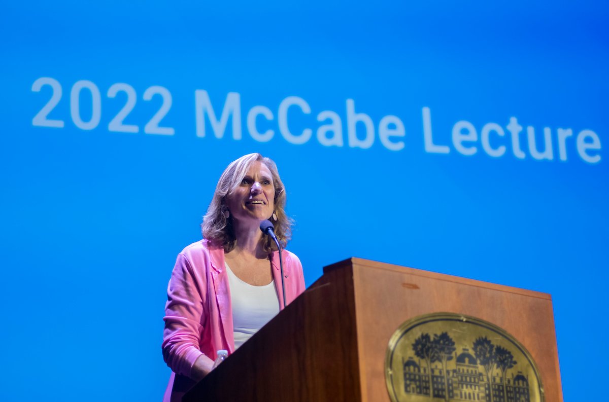 A woman with blonde hair in a pink cardigan stands at a podium in front of a blue screen that says 2022 McCabe Lecture