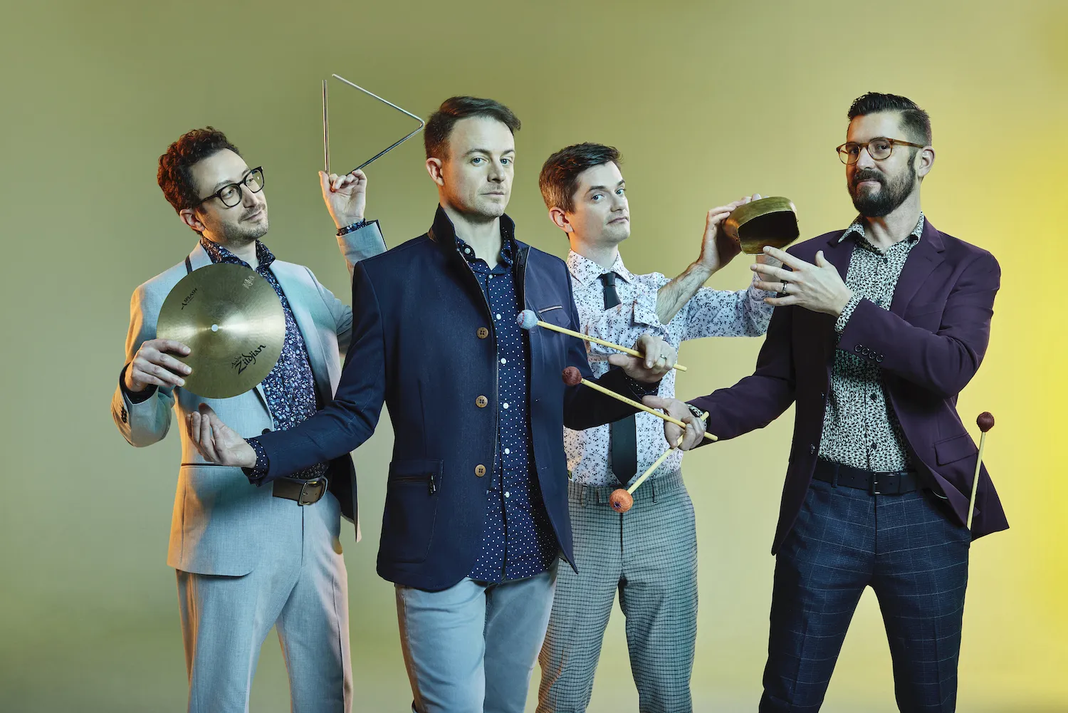 Four musicians holding small percussion instruments standing in front of a green and yellow background