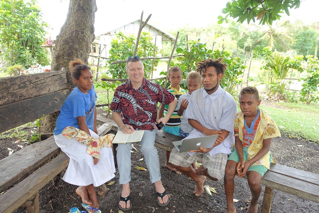 Man sites outside on a bench surrounded by five natives of Vanuatu