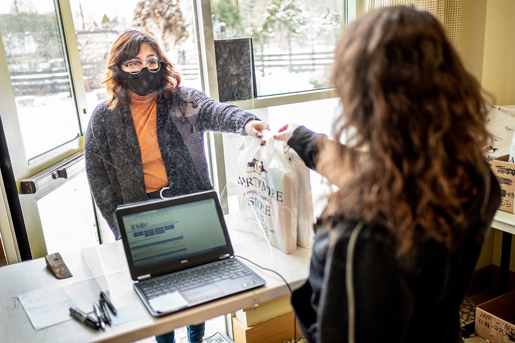 Student in mask handed bag of textbooks from person behind cash register with laptop on desk.