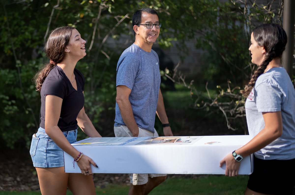 Two young women carry a long rectangular box while an older man assists